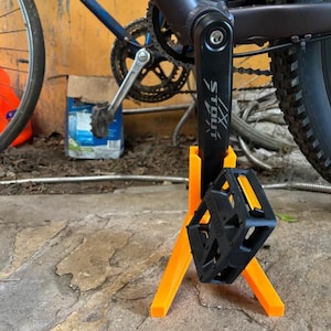 以下が含まれることがあります： 黒い自転車のペダルがオレンジ色のスタンドで支えられています。ペダルは正方形で、表面はテクスチャ加工されています。スタンドはオレンジ色の3本の脚でできています。自転車のフレームは濃いグレーとブルーです。