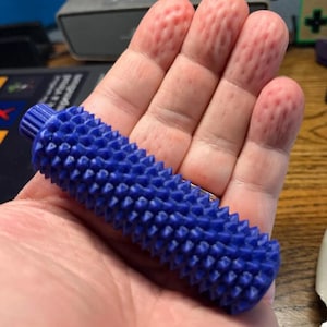 May include: A cylindrical blue object with a textured surface covered in numerous small, pointed spikes. The object features a ribbed cap at one end and a uniform, spiky pattern across its entire length, designed with a tactile, grip-enhancing exterior.