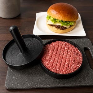 Pode incluir: Uma prensa de hambúrguer de plástico preto com base circular e cabo vertical. A prensa apresenta uma superfície interna texturizada para criar sulcos em hambúrgueres. Um hambúrguer de carne crua com marcas visíveis está na base.