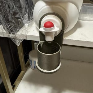May include: A white plastic container with a red and white spout. A gray cylindrical cup is attached below the spout. The container is on a white surface.
