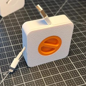 May include: A white square cable winder with a textured surface and a bright orange circular logo featuring a diagonal line. The device has a white charging cable extending from the side and a metallic USB connector protruding from the top edge.