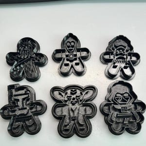 Könnte beinhalten: Sechs schwarze Ausstecher aus Kunststoff in Form von Lebkuchenmännchen, die jeweils so gestaltet sind, dass sie einem Charakter aus einem Science-Fiction-Franchise ähneln. Zu den Charakteren gehören ein Kopfgeldjäger, ein Droide und ein Sith-Lord.