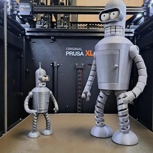 May include: Two gray 3D-printed Bender robot figurines from Futurama, one large and one small, stand side-by-side. The larger Bender has a door on its torso and coiled legs. The background features a 3D printer with the text "ORIGINAL PRUSA XL".