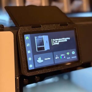 Puede incluir: Una pantalla de control de impresora 3D negra con marco texturizado y tapa abatible. La pantalla muestra un menú con iconos, un gráfico de impresora y el texto: The first rule of 3D printing is...do...talk about 3D printing? Print Files, 41C y Wi-Fi.