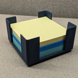 May include: A dark gray square plastic holder containing a stack of square sticky notes. The stack consists of a top layer of pale yellow paper, a middle layer of medium blue paper, and a bottom layer of light teal paper, all neatly aligned.