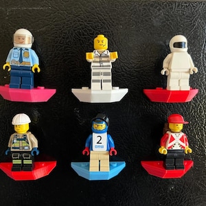 Op de afbeelding: Zes plastic minifiguren gemonteerd op individuele gekleurde voetjes. De figuren omvatten een politieagent, een gevangene in gestreepte kleding, een wit gekleed figuur, een bouwvakker, een blauw figuur met het nummer 2 en een figuur met een rode pet.