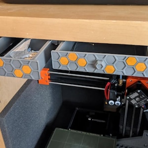 Op de afbeelding: Een set grijze 3D-geprinte opbergladen met een zeshoekig honingraatpatroon op de voorpanelen, voorzien van oranje zeshoekige inzetstukken en een centrale zeshoekige knop. De laden zijn gemonteerd onder een houten oppervlak boven een 3D-printer.