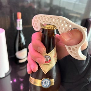 Può includere: Un apribottiglie bianco con un design a nido d'ape apre una bottiglia di birra marrone. La bottiglia ha un'etichetta dorata e bianca. Lo sfondo è sfocato, mostrando una bottiglia con un tappo.