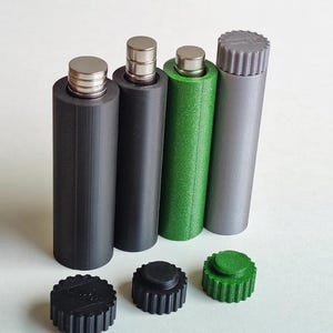 May include: Four cylindrical containers with metallic magnetic tops, including two black, one green, and one grey unit. Three matching circular, ridged screw-on caps are placed in front of the containers, coloured black, black, and green respectively.