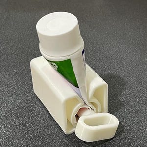 May include: A white plastic toothpaste tube squeezer featuring a rectangular base with a central slot for the tube and a separate, detachable white oval-shaped rolling key used to compress the tube and dispense the contents efficiently.