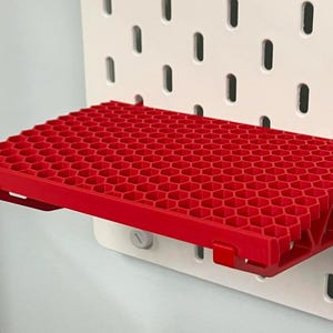 May include: A bright red rectangular shelf featuring a textured honeycomb pattern on its top surface. The shelf is designed with integrated mounting clips that attach directly into the vertical slots of a white pegboard storage panel.