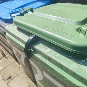 Puede incluir: Un contenedor de basura de plástico verde con un clip de plástico negro que asegura la tapa. La tapa presenta el texto en relieve GEEN GROF AFVAL INWERPEN. Un contenedor de basura de plástico azul es parcialmente visible al fondo.