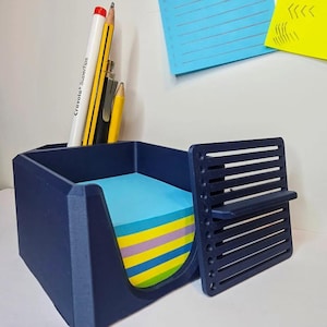 Puede incluir: Un organizador de escritorio azul marino con un compartimento para notas adhesivas y una sección para bolígrafos y lápices. El organizador contiene una pila de notas adhesivas coloridas y varios utensilios de escritura, incluido un rotulador blanco y un lápiz amarillo.