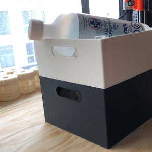 May include: A rectangular storage bin with a white top section and a dark gray bottom section, featuring integrated handles on the sides. A clear plastic bottle of 99% isopropyl alcohol is resting horizontally inside the bin.