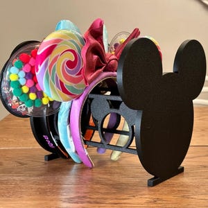 May include: A black acrylic headband display stand shaped like a mouse silhouette, holding multiple headbands. The headbands feature various designs, including a clear ear piece filled with colorful pom-poms and a swirl-patterned lollipop ear design.