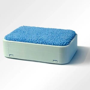 Puede incluir: Un recipiente rectangular con una base de plástico liso en color verde menta claro y una parte superior de tela texturizada en azul brillante. La base presenta pequeños detalles integrados tipo bisagra en el borde inferior.