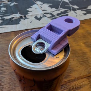 Op de afbeelding: Een paarse 3D-geprinte blikopener bevestigd aan een gedeeltelijk geopende aluminium can. De blikopener heeft een rechthoekig ontwerp met een gat voor een sleutelhanger. De can is oranje met een zilveren bovenkant.