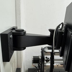 Puede incluir: Un soporte de pared articulado negro para monitor que cuenta con una placa base rectangular fijada a una superficie vertical, un brazo de extensión horizontal con articulación pivotante y un soporte plano unido a la parte trasera de una pantalla negra.