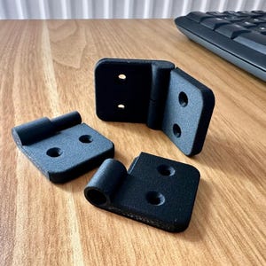 May include: Three matte black hinges with a textured finish. Each hinge features a rectangular plate with two countersunk screw holes and a cylindrical barrel for a pin. The hinges are arranged in a group, showing both flat and folded orientations.