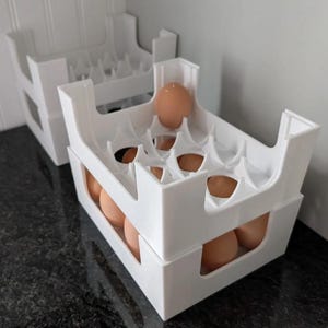 May include: Two white plastic egg storage organizers stacked vertically. Each rectangular holder features a grid of molded circular cutouts designed to securely hold individual brown eggs in place, with open sides for visibility and access.