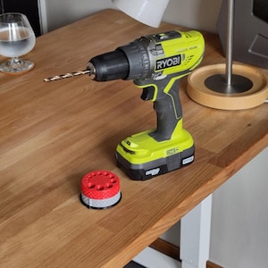May include: A lime green and black Ryobi One+ cordless drill with a metal drill bit inserted into the chuck. Next to the drill sits a small, round, two-tiered accessory holder with a red top section and a grey base, both featuring a textured pattern.