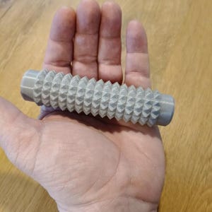 May include: A cylindrical grey massage roller featuring a textured surface with numerous small, pointed, pyramid-shaped protrusions arranged in rows along the entire length of the object, designed for tactile stimulation and pressure application.