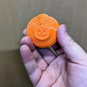 May include: A bright orange, circular, 3D-printed object featuring a concentric, layered design that creates a rippled effect. At the center of the disc is a carved jack-o'-lantern face with triangular eyes and a jagged, smiling mouth.