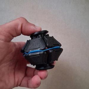 Può includere: Un piccolo giocattolo antistress sferico composto da diversi segmenti in plastica nera stampati in 3D, collegati da un elastico blu brillante. L'oggetto presenta una superficie testurizzata e un'apertura circolare sia sulla parte superiore che su quella inferiore.