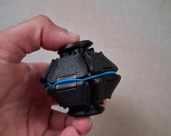 Può includere: Un piccolo giocattolo antistress sferico composto da diversi segmenti in plastica nera stampati in 3D, collegati da un elastico blu brillante. L'oggetto presenta una superficie testurizzata e un'apertura circolare sia sulla parte superiore che su quella inferiore.