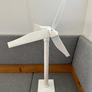 Puede incluir: Un modelo de turbina eólica impreso en 3D de color blanco que presenta una torre cilíndrica vertical, una base cuadrada y un conjunto de rotor de tres palas unido a un cubo central. La estructura es de material blanco mate con líneas.