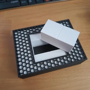 Può includere: Una base rettangolare nera con un motivo a griglia a nido d'ape bianco sulla superficie. Un blocco rettangolare bianco è posizionato diagonalmente al centro della base, coprendo parzialmente un'apertura rettangolare incassata bianca e nera.