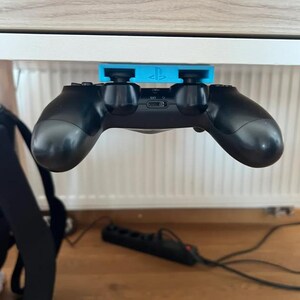 May include: A black PlayStation controller is held in place by a blue holder, mounted under a white cabinet. The holder has the PlayStation logo. The controller is suspended in mid-air, with the buttons and joysticks visible.