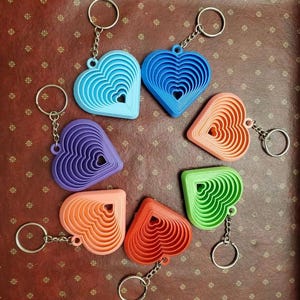 May include: A collection of seven heart-shaped keychains in various colors including light blue, dark blue, peach, green, red, orange, and purple. Each keychain features a concentric, layered design with a central cutout and a metal ring.