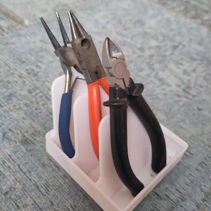 May include: A white plastic organizer stand holding three pairs of pliers. The pliers feature distinct handle colors: one pair with blue grips, one pair with orange grips, and one pair with black grips. The stand has three vertical slots.