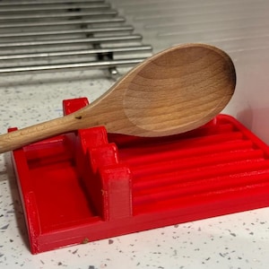 May include: A bright red plastic spoon rest featuring a rectangular base with multiple raised ridges and a notched support structure, holding a single light brown wooden spoon with a smooth, rounded bowl and a long, slender handle.