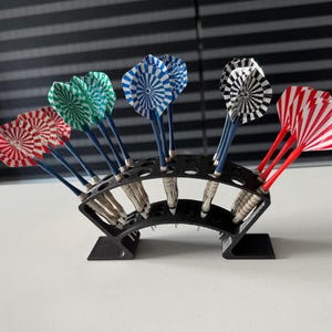 Peut inclure: Un support à fléchettes noir en plastique incurvé contenant douze fléchettes avec des corps métalliques et des ailettes à motifs rouges, verts, bleus, noirs et blancs. Le support présente plusieurs fentes circulaires disposées sur deux rangées.