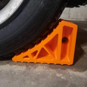 May include: Bright orange wheel chock positioned under a black tire. The chock is made of durable plastic and features a triangular design with cutouts for weight reduction. The tire is part of a vehicle, likely a car or truck.
