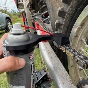 Puede incluir: Un aerosol plateado con un aplicador negro se utiliza para lubricar una cadena de motocicleta. La motocicleta es roja y plateada, con un neumático y una cadena negros. El fondo incluye hierba y un coche.