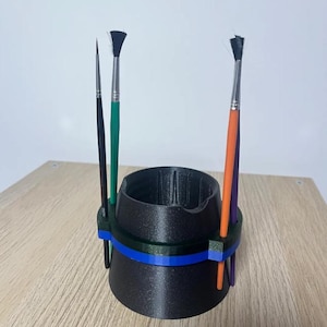 May include: A black conical paint brush holder with a blue and green ring attachment. The holder features four vertical slots containing four paint brushes with black, green, orange, and purple handles, arranged around the central structure.