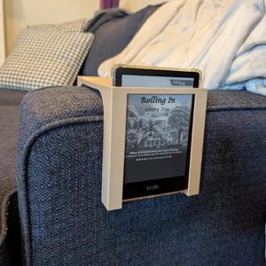 Könnte beinhalten: Ein beigefarbener E-Reader-Halter aus Kunststoff, der an einer Sofa-Armlehne befestigt wird. Der Halter verfügt über einen rechteckigen Rahmen, der einen schwarzen Kindle E-Reader sicher hält, auf dessen Bildschirm das Buchcover Rolling In von Ginny Dye zu sehen ist.