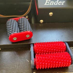 Könnte beinhalten: Zwei manuelle Massageroller mit stacheligen zylindrischen Oberflächen. Ein Roller hat eine schwarze Stacheloberfläche mit roten Endkappen, der andere eine rote Stacheloberfläche mit schwarzen Endkappen, beide mit sichtbaren Metallschrauben.