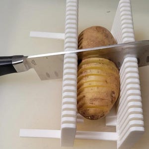 Könnte beinhalten: Eine weiße Kartoffelschneidehilfe aus Kunststoff mit vertikalen Schlitzen auf beiden Seiten. Eine braune Kartoffel liegt zwischen den beiden Führungswänden und ein Küchenmesser aus Edelstahl mit schwarzem Griff steckt in den Schlitzen.