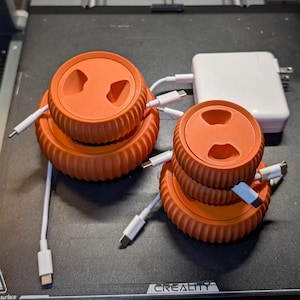 May include: Two orange 3D-printed cable organizers with a ridged design, each with a circular top featuring two openings. White charging cables are inserted into the organizers. A white power adapter is also present.