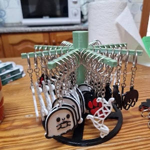 Könnte beinhalten: Ein Präsentationsständer mit verschiedenen Schlüsselanhängern mit unterschiedlichen Designs. Die Schlüsselanhänger zeigen einen weißen Geist, einen Pfotenabdruck und andere schwarz-weiße Formen. Der Ständer ist grün und schwarz, die Schlüsselanhänger sind mit silbernen Ringen befestigt.