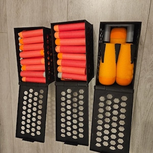 May include: Three black rectangular storage containers with honeycomb-patterned lids. Two containers are filled with rows of cylindrical red foam darts with orange tips. The third container holds three larger orange foam projectiles with black fins.