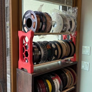 May include: A red 3D printed storage rack with a honeycomb side pattern holds two rows of circular filament spools in various colors. The rack sits atop a dark brown wooden shelf, which also contains a third row of filament spools in multiple colors.