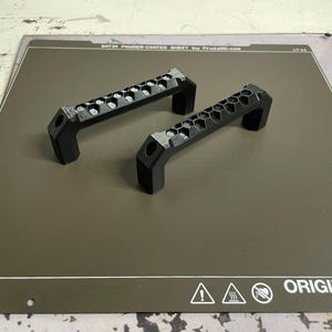 May include: Two black, geometric handles with a honeycomb design. The handles are made of a matte material and are positioned on a gray surface. The handles have a rectangular shape with a curved grip.
