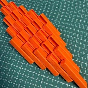 May include: A collection of bright orange plastic clips arranged in a diagonal pattern. The clips are rectangular with a U-shaped opening and a ridged interior. The background is a green cutting mat with a grid pattern.