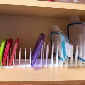 May include: A white plastic lid organizer inside a wooden cabinet. The organizer holds various colorful lids, including green, red, and purple. Several clear and blue food storage containers are also present.