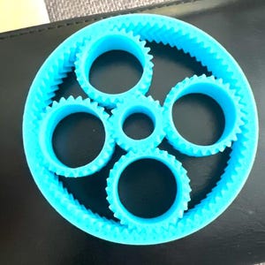 Könnte beinhalten: Ein helles, cyanfarbenes, 3D-gedrucktes Planetengetriebe, bestehend aus einem großen äußeren Hohlrad mit Innenverzahnung, vier kleineren Planetenrädern mit Außenverzahnung und einem zentralen Sonnenrad in einer kreisförmigen Anordnung.